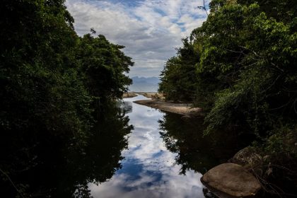 Rio cercado por vegetação exuberante, refletindo o céu nublado em uma paisagem costeira brasileira.