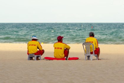 Três guarda-vidas uniformizados sentados na areia, de frente para o mar, durante a vigilância da praia.