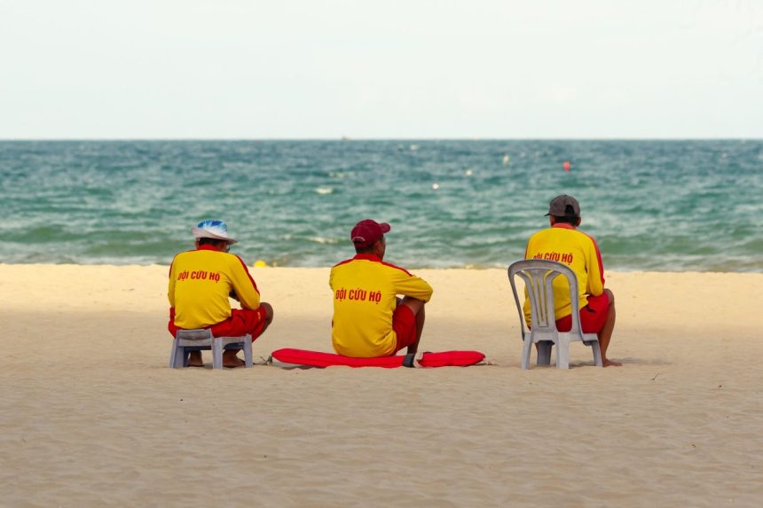 Três guarda-vidas uniformizados sentados na areia, de frente para o mar, durante a vigilância da praia.