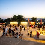Público se reúne ao entardecer em um festival ao ar livre com barracas de comida e clima festivo.